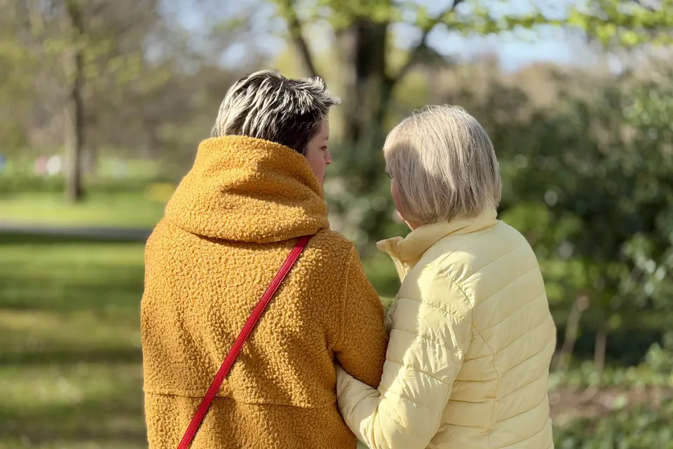 Anja Mattausch und eine Seniorin, von hinten, Arm in Arm im Park